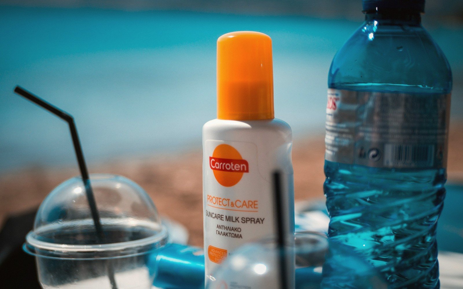 Bottle of Carroten suncare milk spray beside a water bottle and iced drink